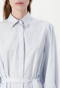 Camicia a righe azzurra chiara con collo, bottoni frontali e vita legata. Presenta maniche lunghe e gonfiate e una texture liscia.