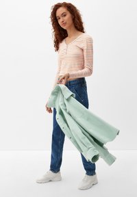Gestreiftes Langarmoberteil in Rosa und Orange, V-Ausschnitt, kombiniert mit blauen Jeans und hält ein hellgrünes Hemd mit Knöpfen. Weiße Sneaker.
