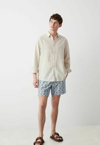 Camicia button-up in lino color beige chiaro, abbinata a pantaloni corti blu con motivi astratti, e sandali scivolati marroni.