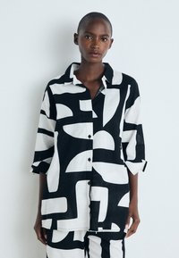 Camisa de patrón en blanco y negro con cierre de botones en la parte delantera y mangas de tres cuartos. La tela parece ligera y texturizada.