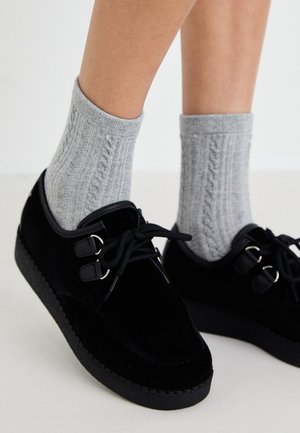 Chaussures en velours noir à bout rond, ornées de deux accents en métal argenté et de lacets noirs, associées à des chaussettes en tricot torsadé grises et texturées.