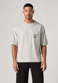 Γκρι βαμβακερό T-shirt με στρογγυλή λαιμόκοψη, κοντά μανίκια και τσέπη στο στήθος με σκούρο μπλε γραφικό τύπωμα. Φορεμένο με σκούρο παντελόνι.