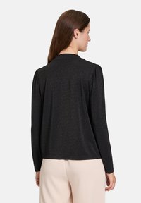 Schwarzes Langarmoberteil mit subtilen Schimmer, Rundhalsausschnitt und gerafften Schultern, kombiniert mit hellen Hosen.