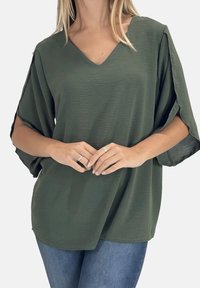 Olijfgroene blouse met V-hals, korte vleermuismouwen, een gestructureerde stof en een losse pasvorm, gecombineerd met lichtblauwe jeans.