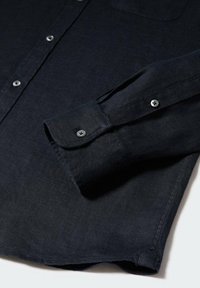 Chemise noire à manches longues avec boutons visibles sur les poignets et la patte de boutonnage avant, posée à plat sur une surface blanche.