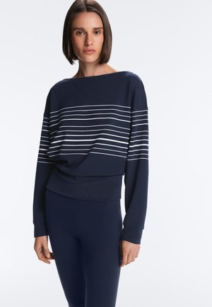 Donna con capelli corti e scuri che indossa una maglia a maniche lunghe blu scuro con strisce orizzontali bianche e pantaloni blu scuro, in piedi davanti a uno sfondo bianco.