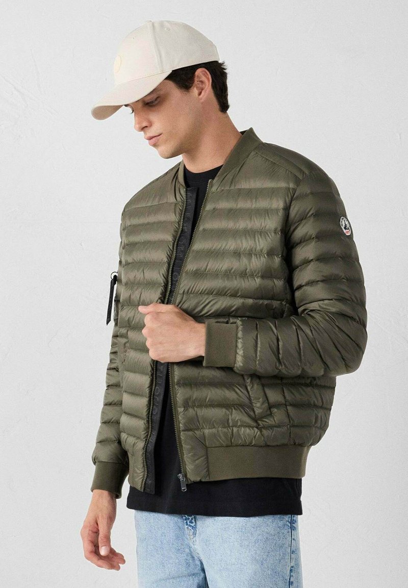Giacca puffer verde oliva con design a cassettoni orizzontali, chiusura con zip, polsini a coste e tasca laterale. Indossata sopra una maglietta nera e jeans chiari.