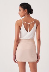 Top bianco senza maniche con scollatura a V profonda sul retro, abbinato a una mini gonna attillata e trasparente di colore beige con un design arricciato.