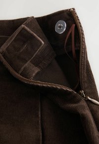 Brown corduroy fabric with a textured surface, featuring a zip closure and a button. Accents include a fabric tag and smooth zipper.