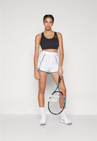Svart sport-BH och vita shorts med turkosa detaljer, med en Lacoste-logotyp. Modellen håller en tenniskö. Skorna är vita sportskor.