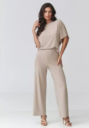 Kobieta z długimi ciemnymi włosami, ubrana w luźną beżową bluzkę, dopasowane szerokie spodnie i beżowe sandały na wysokim obcasie, stojąca na jednolitym tle.