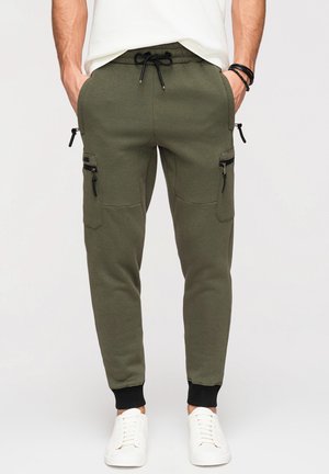 Homme en pantalon de jogging vert olive avec des poches latérales zippées noires, des poignets élastiques et des baskets blanches, mains dans les poches.