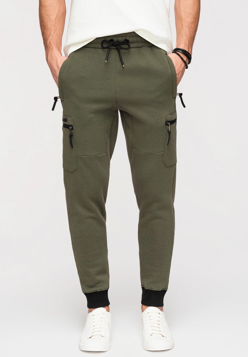 Homme en pantalon de jogging vert olive avec des poches latérales zippées noires, des poignets élastiques et des baskets blanches, mains dans les poches.