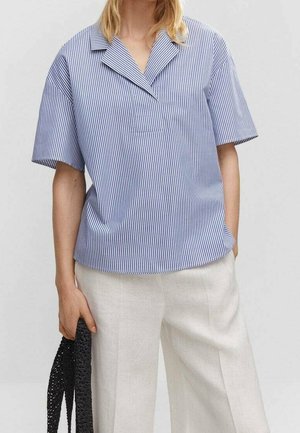 Persoon draagt een blauw-wit verticaal gestreept shirt met korte mouwen en licht beige broek, houdt een zwarte geweven tas in één hand.