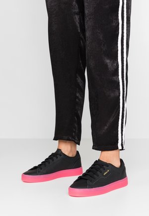 Czarne sneakersy Adidas z jaskraworóżowymi podeszwami, noszone z czarnymi spodniami dresowymi z białymi paskami po bokach.