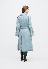 Mulher vista de costas a usar um casaco trench coat de ganga azul claro com cinto e botas pretas de salto, contra um fundo branco.