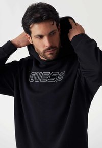 Felpa nera con cappuccio realizzata in tessuto morbido, dotata di una tasca a marsupio frontale e caratterizzata da un logo bianco "GUESS" in evidenza sul petto.