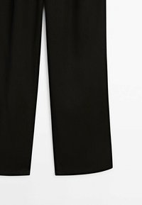 Pantalon noir à jambe droite suspendu sur un fond blanc, montrant la moitié inférieure à partir des genoux.