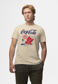 Mladý muž v béžovém tričku Coca-Cola s červenou plechovkou a logem v retro stylu, stojící s jednou rukou v kapse na bílém pozadí.