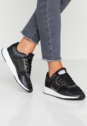 Personne portant des baskets noires et grises avec des semelles blanches et un jean gris coupé avec des ourlets effilochés, debout sur une surface lisse.