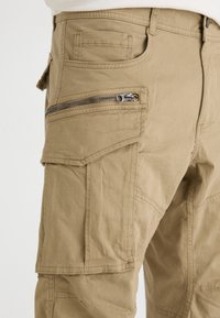 Beige cargobroek met meerdere zakken, een ritszak aan de zijkant en zichtbare stiksels. Gemaakt van een duurzame katoenen stof.