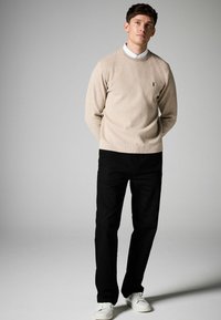 Beige striktrøje med rund halsudskæring og broderet logo, parret med sorte lige bukser og hvide sneakers, på en neutral baggrund.