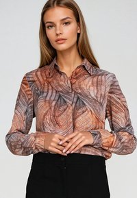 Młoda kobieta z długimi blond włosami, ubrana w wzorzystą brązowo-szaro button-up bluzkę i czarne spodnie, z rękami złożonymi na talii.