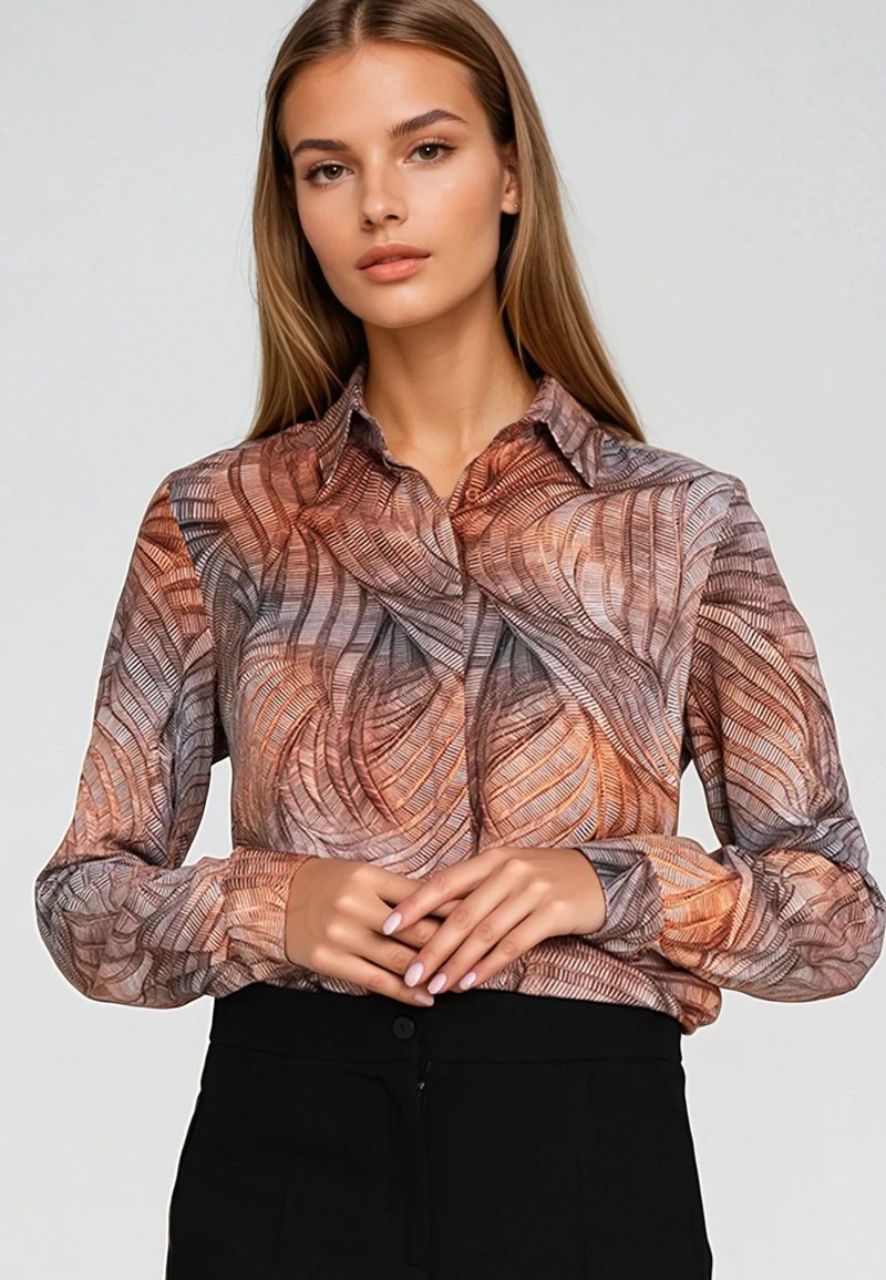 Młoda kobieta z długimi blond włosami, ubrana w wzorzystą brązowo-szaro button-up bluzkę i czarne spodnie, z rękami złożonymi na talii.