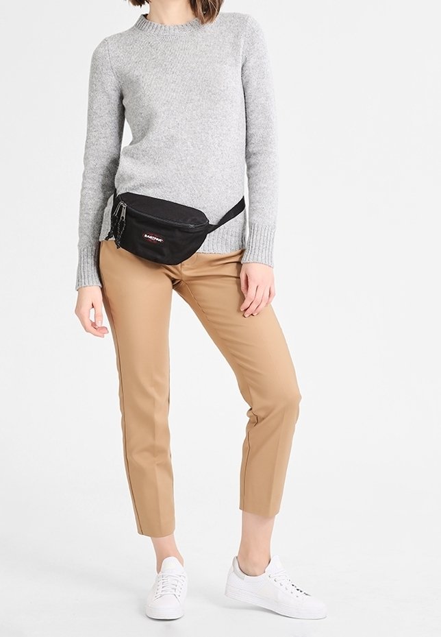 Personne portant un pull gris, un pantalon beige court, des baskets blanches et un sac banane noir avec une poche zippée à l'avant.