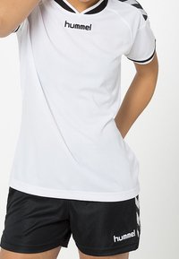 Persona vestida con una camiseta deportiva blanca de hummel y pantalones cortos negros de hummel, de pie con un brazo doblado y la otra mano en el bolsillo.
