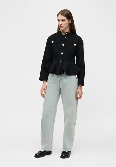 Γυναίκα που στέκεται φορώντας μαύρο σακάκι με κουμπιά και πεπλουμ, ανοιχτό γκρι τζιν ίσιας γραμμής και μαύρα μοκασίνια, μπροστά σε μια απλή ανοιχτόχρωμη φόντο.