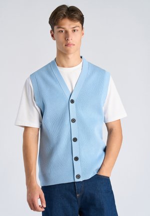 Ung mand iført en lyseblå knappet strikvest over en hvid t-shirt og mørkeblå jeans, stående med den ene hånd i lommen.