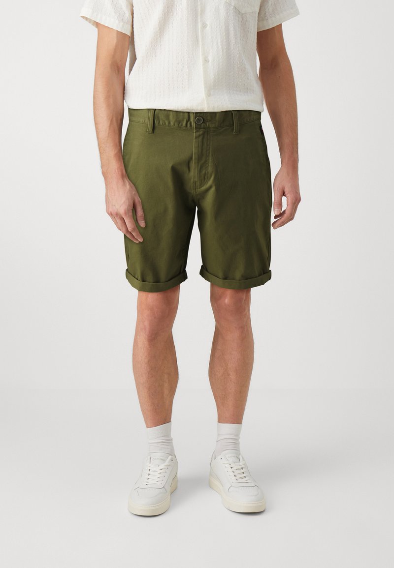 Pantaloni corti verde oliva con taglio dritto, risvolti e chiusura con bottone frontale. Il tessuto è leggero e dalla consistenza liscia.