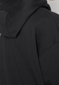 Svart hoodie i mjukt material, med en stor huva och sömlös design. Texturen framstår som slät med minimala sömnadsdetaljer.