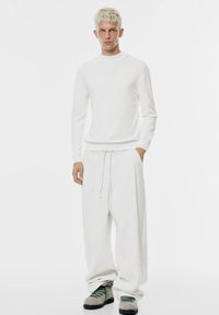 Weißes, gestricktes Langarmshirt kombiniert mit locker sitzenden, hellgrauen Jogginghosen. Die Schuhe haben eine klobige Sohle und grüne Schnürsenkel.