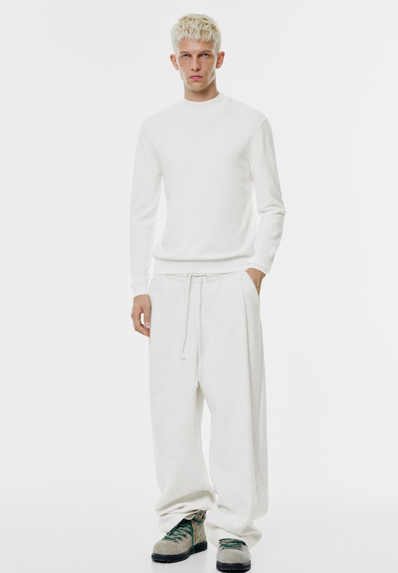 Weißes, gestricktes Langarmshirt kombiniert mit locker sitzenden, hellgrauen Jogginghosen. Die Schuhe haben eine klobige Sohle und grüne Schnürsenkel.