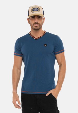 Homme portant une casquette beige et marine, un t-shirt bleu à manches courtes avec col en V et bordure rouge, et un pantalon noir, debout avec une main dans la poche.