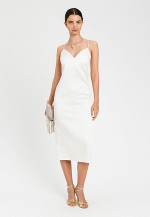 Donna che indossa un abito midi bianco senza maniche con spalline sottili, tacchi dorati con cinturini, una collana con ciondolo e tiene una pochette con frange di perline.