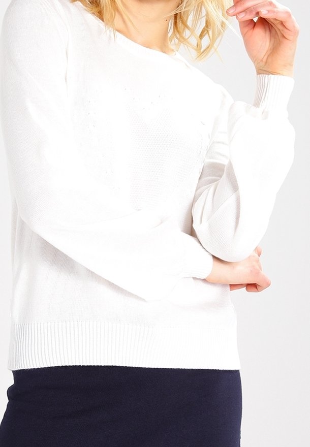 Femme portant un pull en maille texturée blanche à manches longues, associée à une jupe sombre, main près du visage et bras croisés.