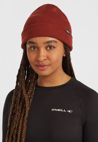 Gorro de punto de canalé rojo con un ajuste ceñido, que presenta una pequeña etiqueta negra de O'Neill; llevado con una camiseta de manga larga negra ajustada.