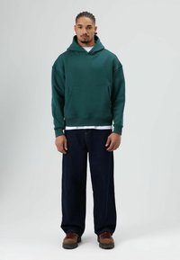 Sweat à capuche vert avec poche avant, porté sur une chemise blanche, associé à un jean ample foncé et des chaussures marron à lacets.