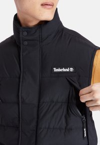 Czarna ocieplana kamizelka z wysokim kołnierzem, z zamykaną na zamek kieszenią i naszywką z logo Timberland. Materiał boczny jest w kolorze piaskowym.