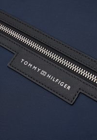 Gros plan sur un tissu bleu marine avec une fermeture éclair noire et une étiquette cousue portant la mention "Tommy Hilfiger" en lettres blanches.