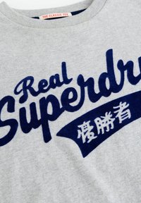 Pilkas medvilninis marškinėliai su tamsiai mėlynu tekstu "Real Superdry" ir papildomomis grafika. Raukšlėta apykaklė ir švelni tekstūra.