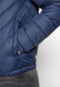Granatowa puchowa kurtka z pikowaniem w kształcie diamentów, elastycznymi mankietami i boczną kieszenią. Gładki, błyszczący materiał o lekkim, ocieplanym charakterze.