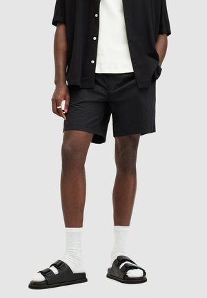 Zwarte shorts met een ontspannen pasvorm, gecombineerd met een wit geribbeld shirt en zwarte sandalen. Katoenen stof, minimalistisch ontwerp en bicolor banden op de sandalen.