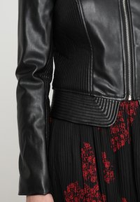 Czarna skórzana kurtka o prążkowanej fakturze i dopasowanej talii. Wyposażona w srebrny zamek błyskawiczny i eleganckie, długie rękawy. Połączona z czarną spódnicą w kwiaty.