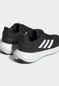 Zapatillas deportivas negras con una parte superior de malla texturizada, diseño de tres rayas blancas y una suela blanca gruesa con una ligera curvatura.