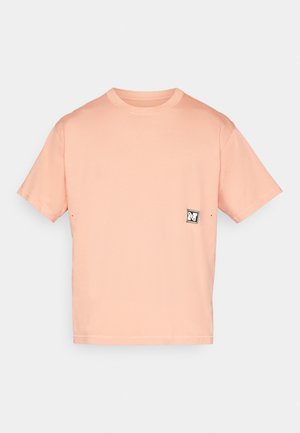 T-shirt à manches courtes et col rond en tissu doux de couleur pêche avec une texture subtile et un petit patch logo noir et blanc sur la poitrine.