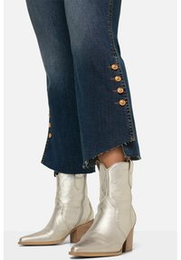 Jeans acampanados de mezclilla azul oscuro con detalles de botones dorados en los costados y dobladillo deshilachado, combinados con botas de vaquero metálicas plateadas y tacón apilado.